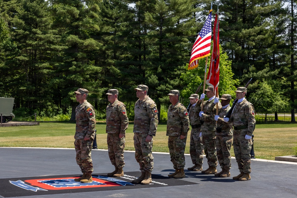 A military color guard ceremony is taking place outdoors on a paved surface surrounded by green trees and grass. Several U.S. Army soldiers in OCP (Operational Camouflage Pattern) uniforms are standing at attention, with two soldiers holding the U.S. flag and a unit guidon/colors. The formation appears to be part of a formal military ceremony, likely a change of command, activation, or similar official event.