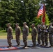 A military color guard ceremony is taking place outdoors on a paved surface surrounded by green trees and grass. Several U.S. Army soldiers in OCP (Operational Camouflage Pattern) uniforms are standing at attention, with two soldiers holding the U.S. flag and a unit guidon/colors. The formation appears to be part of a formal military ceremony, likely a change of command, activation, or similar official event.