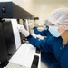 Two workers in a pharmaceutical or laboratory clean-room environment are performing visual inspection of small vials against an illuminated black-and-white background panel. They are wearing full personal protective equipment including hairnets, face masks, safety glasses, gloves, and blue lab coats. Multiple small white-capped vials are visible in a tray in the foreground, consistent with pharmaceutical quality control inspection.