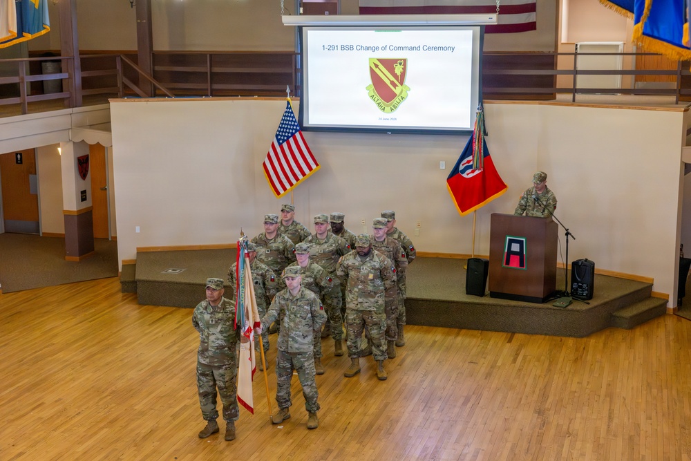 This image depicts a 1-291 BSB (Brigade Support Battalion) Change of Command Ceremony, as indicated by the presentation slide projected on the screen. A formation of soldiers in Army Combat Uniforms (ACU/OCP) stands on and in front of a raised platform, with one soldier at a podium delivering remarks. The ceremony is held indoors in what appears to be a military community or event hall.