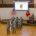 This image depicts a 1-291 BSB (Brigade Support Battalion) Change of Command Ceremony, as indicated by the presentation slide projected on the screen. A formation of soldiers in Army Combat Uniforms (ACU/OCP) stands on and in front of a raised platform, with one soldier at a podium delivering remarks. The ceremony is held indoors in what appears to be a military community or event hall.