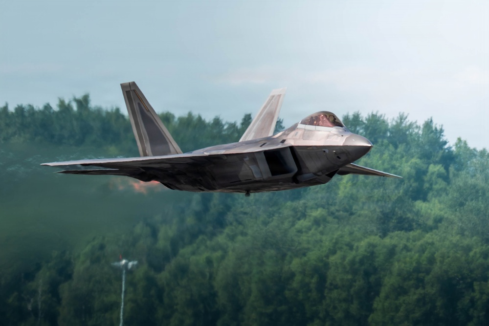 A fifth-generation stealth fighter jet, consistent with the F-22 Raptor design, is captured in low-altitude flight against a backdrop of dense green forest. The aircraft appears to be performing a high-performance maneuver, possibly during an airshow or training exercise. Engine exhaust heat distortion is visible near the rear of the aircraft.