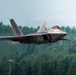 A fifth-generation stealth fighter jet, consistent with the F-22 Raptor design, is captured in low-altitude flight against a backdrop of dense green forest. The aircraft appears to be performing a high-performance maneuver, possibly during an airshow or training exercise. Engine exhaust heat distortion is visible near the rear of the aircraft.