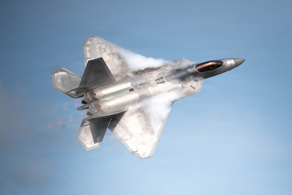 A U.S. Air Force F-22 Raptor fifth-generation stealth fighter jet is captured in flight against a clear blue sky. The aircraft is performing a high-performance maneuver, generating visible vapor condensation clouds across its fuselage and wings due to rapid pressure changes. The weapons bay doors appear to be open, revealing internal payload.