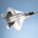 A U.S. Air Force F-22 Raptor fifth-generation stealth fighter jet is captured in flight against a clear blue sky. The aircraft is performing a high-performance maneuver, generating visible vapor condensation clouds across its fuselage and wings due to rapid pressure changes. The weapons bay doors appear to be open, revealing internal payload.