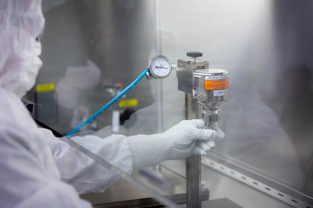 A technician in full cleanroom protective gear (white coverall, hood, and gloves) is operating a pressurized dispensing or filtration device mounted on a metal stand inside what appears to be a cleanroom or laminar flow hood environment. The equipment includes a stainless steel canister labeled 'Power-Crimp' with an orange WARNING label, connected to a blue pressure line and an analog pressure gauge. The setting suggests pharmaceutical, semiconductor, or precision manufacturing operations.
