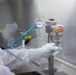 A technician in full cleanroom protective gear (white coverall, hood, and gloves) is operating a pressurized dispensing or filtration device mounted on a metal stand inside what appears to be a cleanroom or laminar flow hood environment. The equipment includes a stainless steel canister labeled 'Power-Crimp' with an orange WARNING label, connected to a blue pressure line and an analog pressure gauge. The setting suggests pharmaceutical, semiconductor, or precision manufacturing operations.