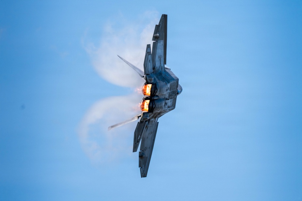 A military stealth fighter jet, consistent with an F-22 Raptor, is photographed from below in a near-vertical banking maneuver against a clear blue sky. Both afterburners are visibly ignited, producing bright orange-yellow exhaust flames. Condensation vapor trails are forming dramatically around the airframe due to low-pressure aerodynamic effects.