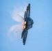 A military stealth fighter jet, consistent with an F-22 Raptor, is photographed from below in a near-vertical banking maneuver against a clear blue sky. Both afterburners are visibly ignited, producing bright orange-yellow exhaust flames. Condensation vapor trails are forming dramatically around the airframe due to low-pressure aerodynamic effects.
