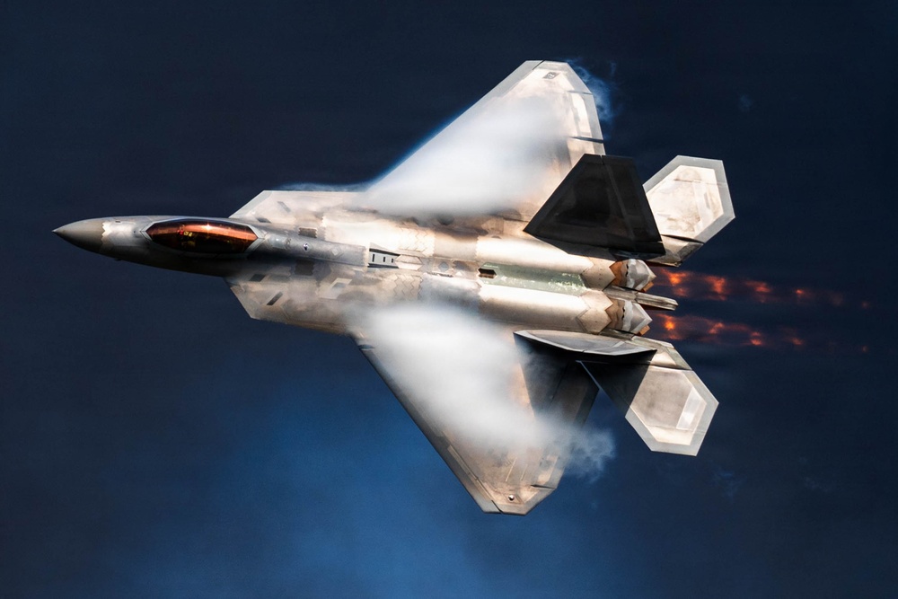 A high-performance stealth fighter jet, identifiable as an F-22 Raptor, is captured in a dramatic low-altitude maneuver against a dark blue sky. The aircraft is photographed from above at an angle, with afterburners ignited and visible vapor condensation forming around the fuselage, indicating high-speed, high-G flight. The image showcases the aircraft's distinctive angular stealth geometry and twin engine exhaust plumes with visible flame.