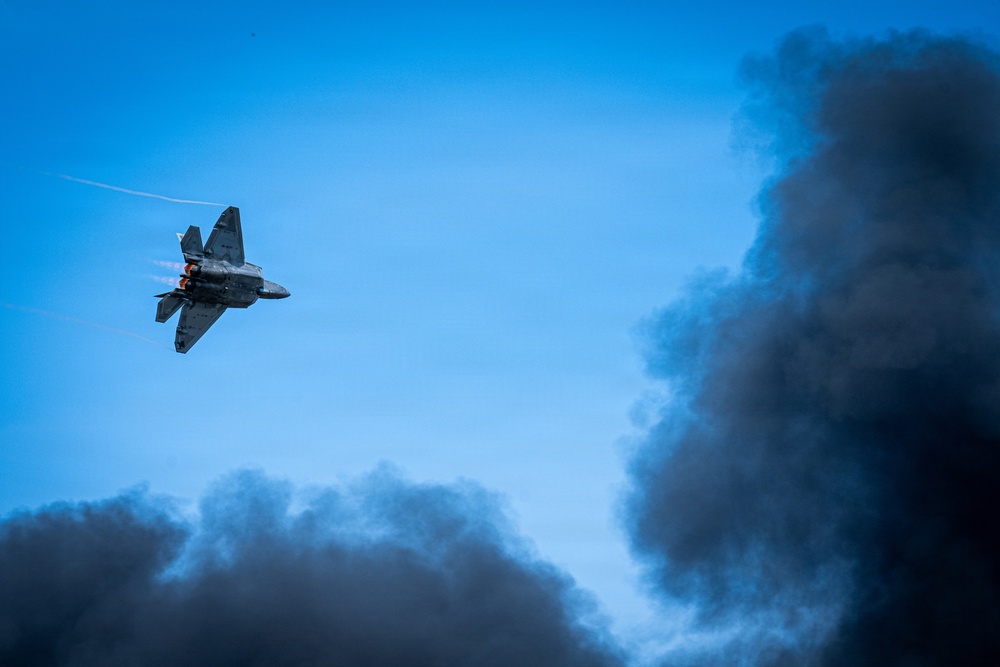 A military jet aircraft, consistent with an F-22 Raptor stealth fighter, is captured in a steep banking maneuver against a clear blue sky. The aircraft's engines appear to be in afterburner, with visible flame exhaust. Large plumes of dark black smoke dominate the lower and right portions of the frame, likely from a ground-based pyrotechnic display or fire, suggesting an airshow demonstration context.