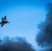 A military jet aircraft, consistent with an F-22 Raptor stealth fighter, is captured in a steep banking maneuver against a clear blue sky. The aircraft's engines appear to be in afterburner, with visible flame exhaust. Large plumes of dark black smoke dominate the lower and right portions of the frame, likely from a ground-based pyrotechnic display or fire, suggesting an airshow demonstration context.