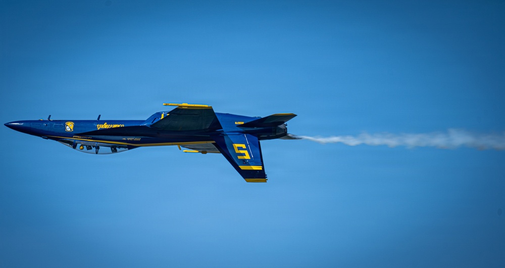 A U.S. Navy Blue Angels F/A-18 Hornet jet, numbered '5', is captured in flight against a clear blue sky. The aircraft is painted in the iconic blue and gold color scheme and is emitting a white smoke trail from its engine exhaust. The jet appears to be performing an aerobatic maneuver, possibly inverted or in a banked turn.