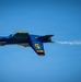 A U.S. Navy Blue Angels F/A-18 Hornet jet, numbered '5', is captured in flight against a clear blue sky. The aircraft is painted in the iconic blue and gold color scheme and is emitting a white smoke trail from its engine exhaust. The jet appears to be performing an aerobatic maneuver, possibly inverted or in a banked turn.