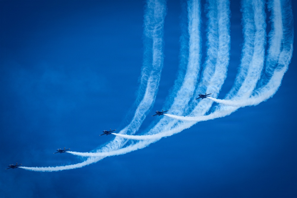 A formation of aerobatic jet aircraft performing a synchronized maneuver against a vivid blue sky. The aircraft are executing what appears to be a break or fan maneuver, with each plane producing a distinct white smoke trail. The trails curve dramatically upward and spread apart, creating a visually striking pattern.