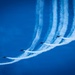 A formation of aerobatic jet aircraft performing a synchronized maneuver against a vivid blue sky. The aircraft are executing what appears to be a break or fan maneuver, with each plane producing a distinct white smoke trail. The trails curve dramatically upward and spread apart, creating a visually striking pattern.
