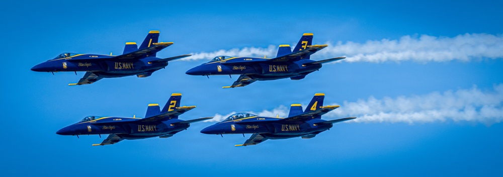 Four U.S. Navy Blue Angels aircraft are flying in a tight diamond formation against a clear blue sky. The jets are numbered 1 through 4 and are producing white smoke trails as they perform their precision aerobatic maneuver. The aircraft are F/A-18 Hornets painted in the iconic blue and gold Blue Angels livery.