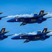 Four U.S. Navy Blue Angels aircraft are flying in a tight diamond formation against a clear blue sky. The jets are numbered 1 through 4 and are producing white smoke trails as they perform their precision aerobatic maneuver. The aircraft are F/A-18 Hornets painted in the iconic blue and gold Blue Angels livery.