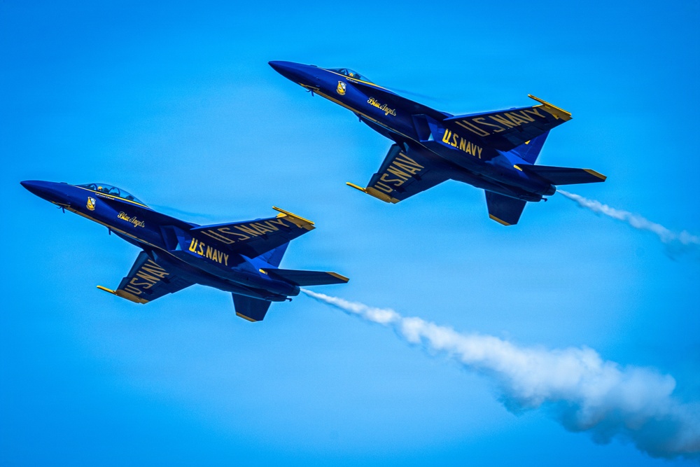 Two U.S. Navy Blue Angels F/A-18 Hornet jets are captured in close formation flight against a clear blue sky. Both aircraft display the iconic dark blue and gold color scheme with 'U.S. NAVY' and 'Blue Angels' markings clearly visible. One or both jets are emitting white smoke trails, characteristic of Blue Angels airshow performances.