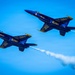 Two U.S. Navy Blue Angels F/A-18 Hornet jets are captured in close formation flight against a clear blue sky. Both aircraft display the iconic dark blue and gold color scheme with 'U.S. NAVY' and 'Blue Angels' markings clearly visible. One or both jets are emitting white smoke trails, characteristic of Blue Angels airshow performances.