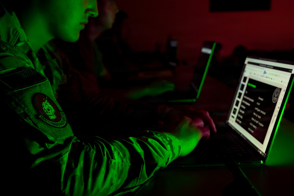 A person in military uniform is working on a laptop in a darkened room illuminated by green light. A second individual is visible in the background also working at a computer. The scene suggests a military cyber operations or intelligence environment.