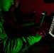 A person in military uniform is working on a laptop in a darkened room illuminated by green light. A second individual is visible in the background also working at a computer. The scene suggests a military cyber operations or intelligence environment.