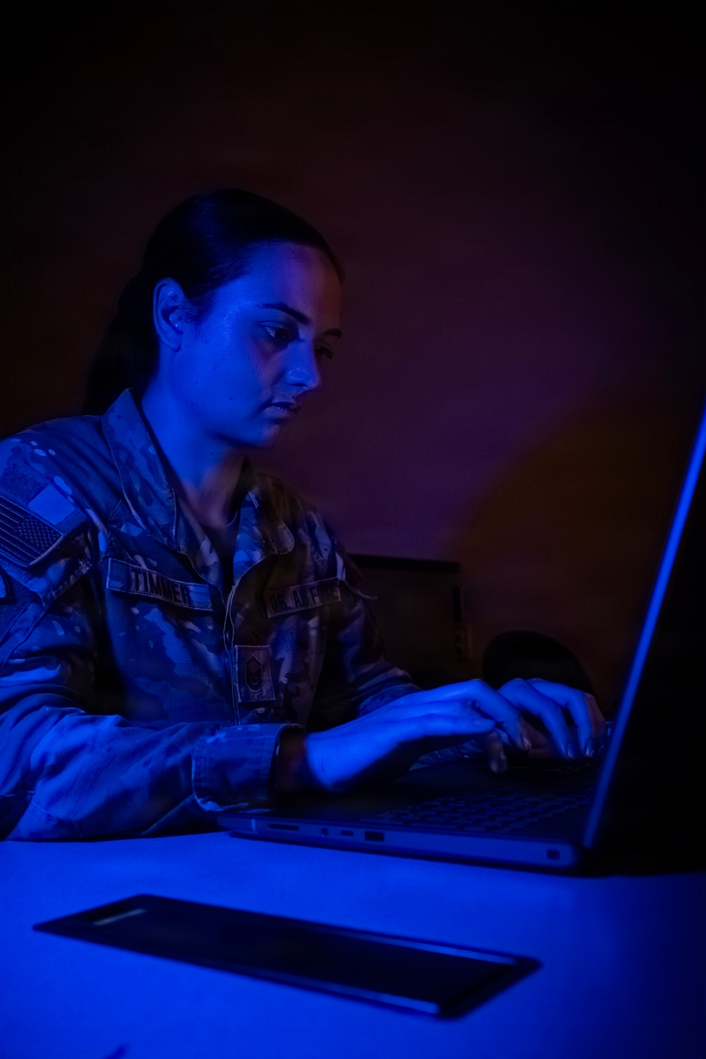 A female military service member in U.S. Air Force OCP (Operational Camouflage Pattern) uniform is working intently on a laptop in a darkened room illuminated by blue light. Her name tape reads 'TIMMER' and the branch tape reads 'US. AIR F[ORCE]'. The dramatic blue lighting and dark background suggest a tactical operations or cyber operations environment.