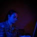 A female military service member in U.S. Air Force OCP (Operational Camouflage Pattern) uniform is working intently on a laptop in a darkened room illuminated by blue light. Her name tape reads 'TIMMER' and the branch tape reads 'US. AIR F[ORCE]'. The dramatic blue lighting and dark background suggest a tactical operations or cyber operations environment.