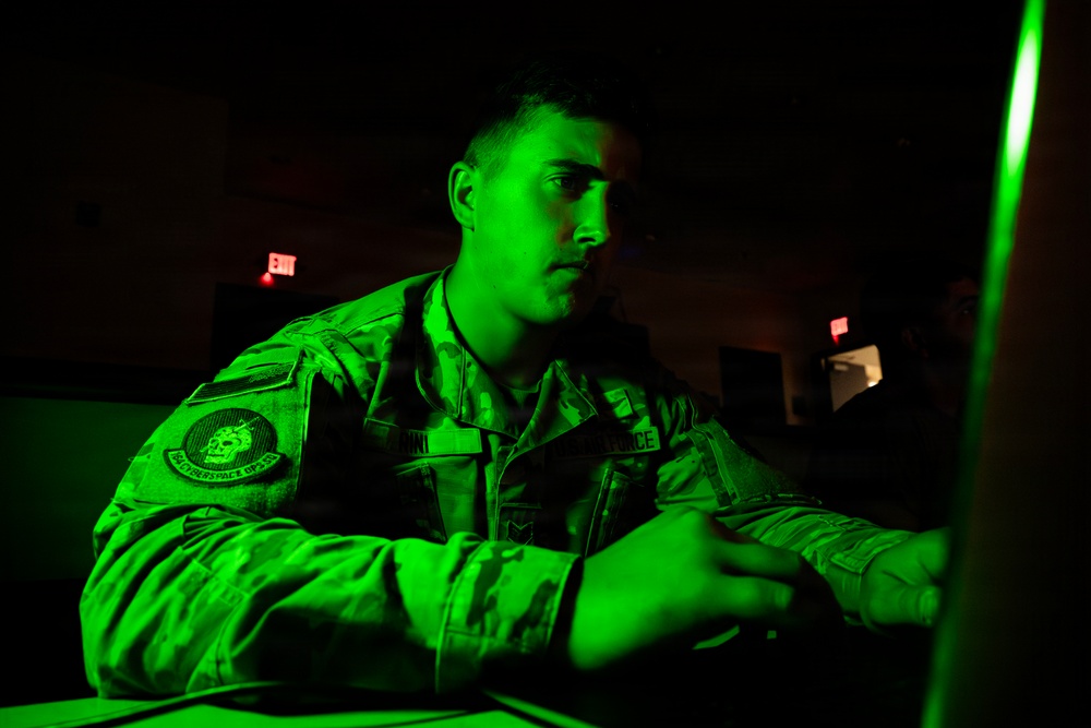 A military service member in camouflage uniform is illuminated by a green monitor glow in a darkened operations room. The individual appears to be working at a computer workstation, focused on a screen out of frame. The setting suggests a cyber operations or command and control environment.