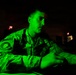 A military service member in camouflage uniform is illuminated by a green monitor glow in a darkened operations room. The individual appears to be working at a computer workstation, focused on a screen out of frame. The setting suggests a cyber operations or command and control environment.