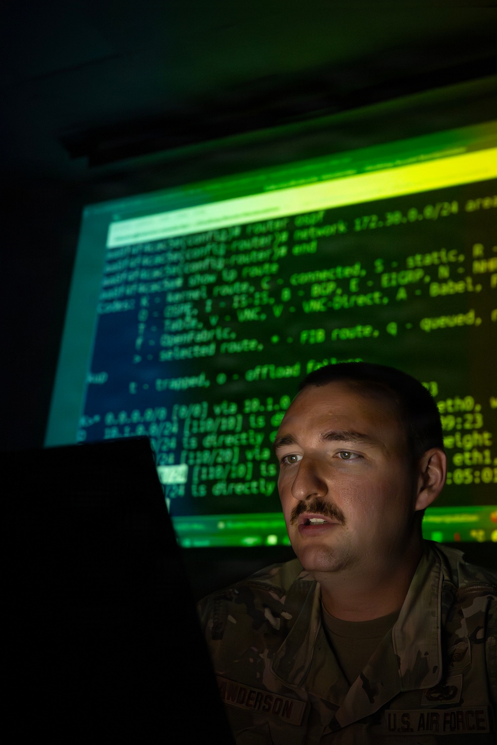 A U.S. Air Force service member is seated in a darkened room, illuminated by the green glow of a large projected screen displaying network routing command-line interface output. The individual is wearing a camouflage uniform with a name tape partially visible reading 'NDERSON' and a 'U.S. AIR F[ORCE]' tape. The dramatic low-key lighting and terminal content suggest a cybersecurity or network operations context.