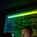 A U.S. Air Force service member is seated in a darkened room, illuminated by the green glow of a large projected screen displaying network routing command-line interface output. The individual is wearing a camouflage uniform with a name tape partially visible reading 'NDERSON' and a 'U.S. AIR F[ORCE]' tape. The dramatic low-key lighting and terminal content suggest a cybersecurity or network operations context.