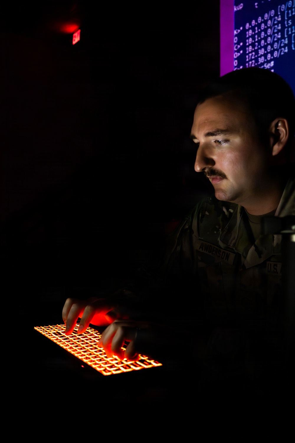 A U.S. military service member, identified by a name tape reading 'ANDERSON' and 'U.S.' on an olive drab uniform, is working at a computer workstation in a darkened room. The individual is typing on a red/orange backlit keyboard while focused on screens displaying what appear to be network routing tables or IP address data. The dramatic low-light environment is illuminated primarily by the keyboard glow, a red exit sign, and blue/purple monitor light.
