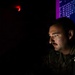 A U.S. military service member, identified by a name tape reading 'ANDERSON' and 'U.S.' on an olive drab uniform, is working at a computer workstation in a darkened room. The individual is typing on a red/orange backlit keyboard while focused on screens displaying what appear to be network routing tables or IP address data. The dramatic low-light environment is illuminated primarily by the keyboard glow, a red exit sign, and blue/purple monitor light.