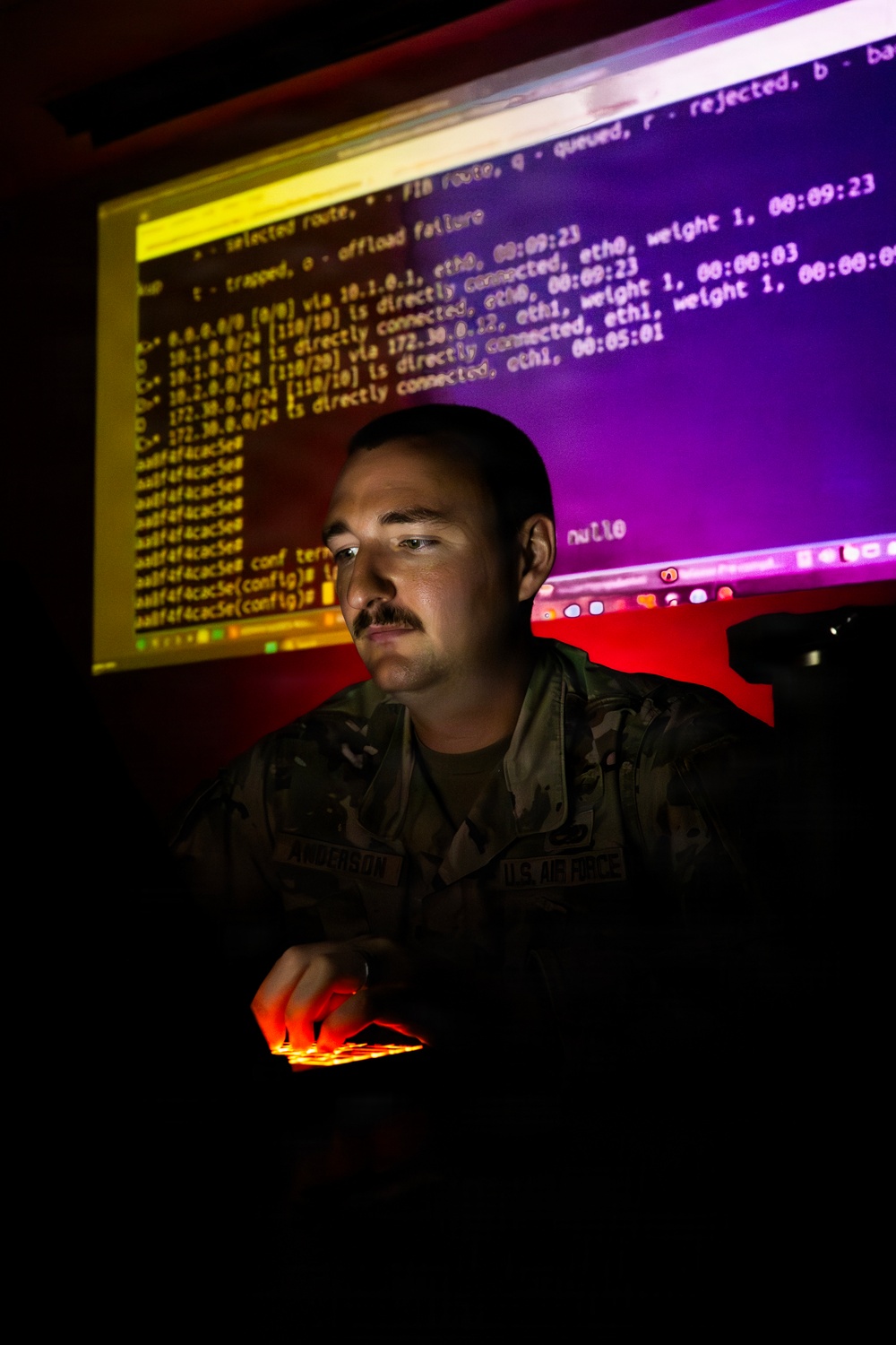 A U.S. Air Force service member, identified by name tape as 'Anderson,' is working at a computer workstation in a darkened room. The individual is illuminated by the glow of large monitors displaying network routing and command-line interface (CLI) output, suggesting cybersecurity or network operations work. The dramatic lighting creates a high-contrast, atmospheric composition typical of military public affairs photography.