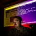 A U.S. Air Force service member, identified by name tape as 'Anderson,' is working at a computer workstation in a darkened room. The individual is illuminated by the glow of large monitors displaying network routing and command-line interface (CLI) output, suggesting cybersecurity or network operations work. The dramatic lighting creates a high-contrast, atmospheric composition typical of military public affairs photography.
