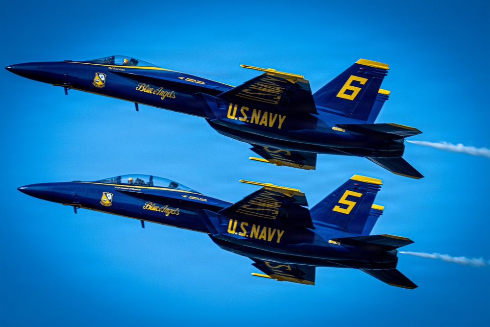 Two U.S. Navy Blue Angels F/A-18 Hornet jets flying in close formation against a clear blue sky. The aircraft are numbered 5 and 6, painted in the iconic blue and gold color scheme with 'Blue Angels' and 'U.S. Navy' markings clearly visible. Both jets are producing white contrails from their engines during what appears to be an airshow performance.