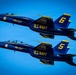 Two U.S. Navy Blue Angels F/A-18 Hornet jets flying in close formation against a clear blue sky. The aircraft are numbered 5 and 6, painted in the iconic blue and gold color scheme with 'Blue Angels' and 'U.S. Navy' markings clearly visible. Both jets are producing white contrails from their engines during what appears to be an airshow performance.
