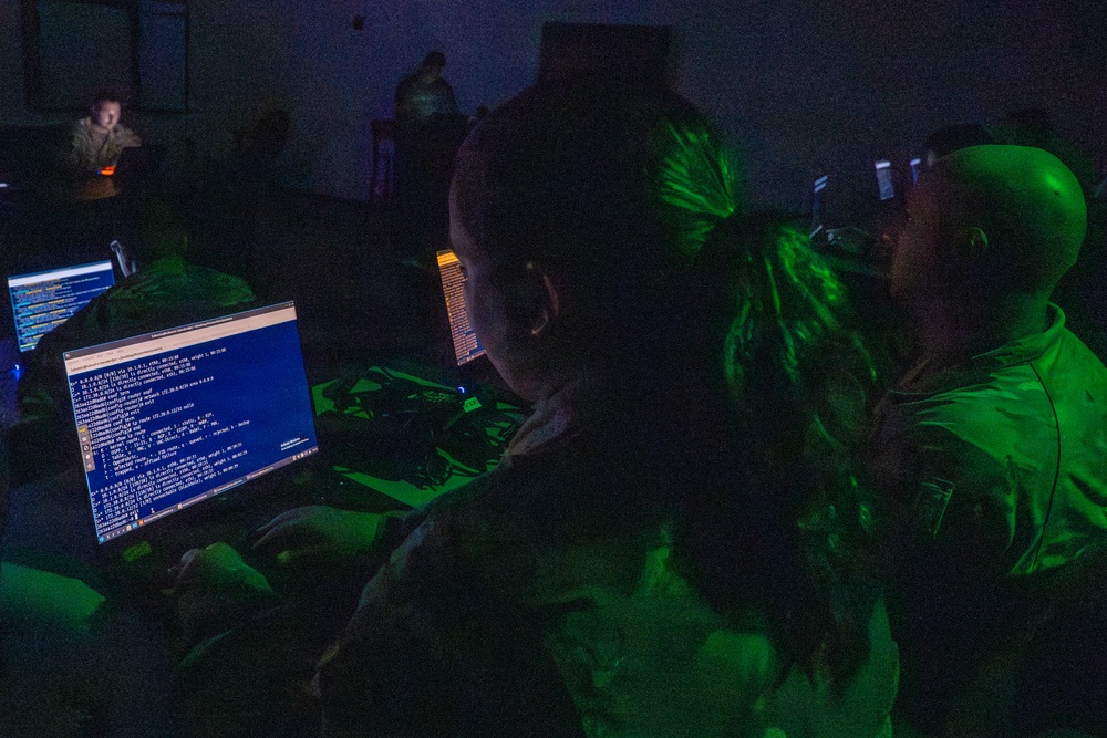 A darkened room with multiple individuals working at computer terminals displaying command-line interfaces and code. The scene is illuminated primarily by monitor glow, with a distinctive green light casting on figures in the foreground. The setting resembles a cybersecurity operations center, training exercise, or capture-the-flag (CTF) competition.