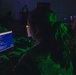 A darkened room with multiple individuals working at computer terminals displaying command-line interfaces and code. The scene is illuminated primarily by monitor glow, with a distinctive green light casting on figures in the foreground. The setting resembles a cybersecurity operations center, training exercise, or capture-the-flag (CTF) competition.