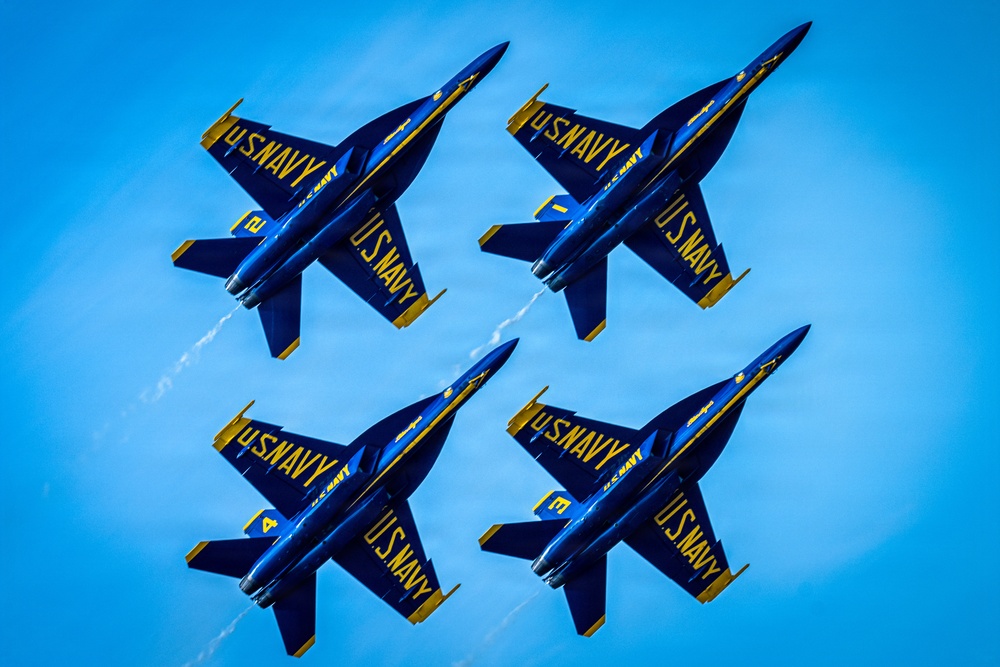 Four U.S. Navy Blue Angels F/A-18 Hornet jets fly in a tight diamond formation against a clear blue sky. The aircraft are painted in their iconic blue and gold livery with 'U.S. NAVY' lettering prominently displayed on the undersides. At least one aircraft is producing a visible white vapor/smoke trail.