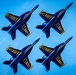 Four U.S. Navy Blue Angels F/A-18 Hornet jets fly in a tight diamond formation against a clear blue sky. The aircraft are painted in their iconic blue and gold livery with 'U.S. NAVY' lettering prominently displayed on the undersides. At least one aircraft is producing a visible white vapor/smoke trail.