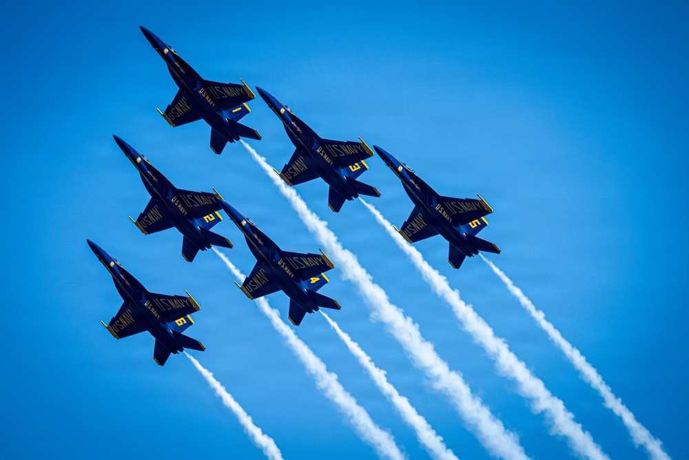 Six U.S. Navy Blue Angels aircraft fly in a tight diamond formation against a clear blue sky, each trailing white smoke. The jets are numbered 1 through 6 and are painted in the iconic navy blue and gold color scheme. This appears to be a precision aerobatic demonstration, likely at an air show.