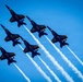 Six U.S. Navy Blue Angels aircraft fly in a tight diamond formation against a clear blue sky, each trailing white smoke. The jets are numbered 1 through 6 and are painted in the iconic navy blue and gold color scheme. This appears to be a precision aerobatic demonstration, likely at an air show.