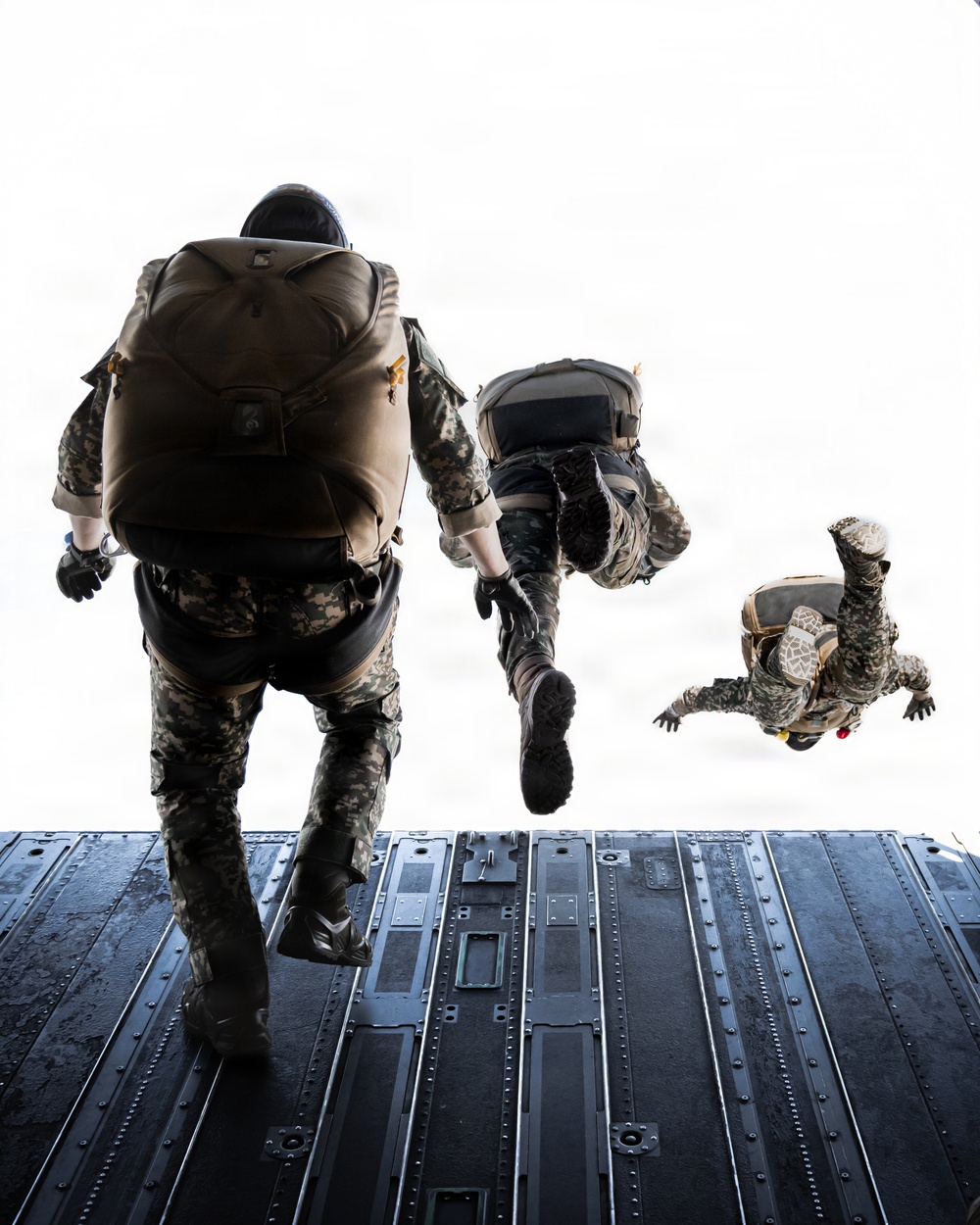 Three military personnel are conducting a parachute jump from the open rear ramp of a military cargo aircraft. Two jumpers are still on the ramp in the process of exiting, while a third is already airborne and visible in freefall position. All individuals are wearing camouflage uniforms and carrying parachute packs.