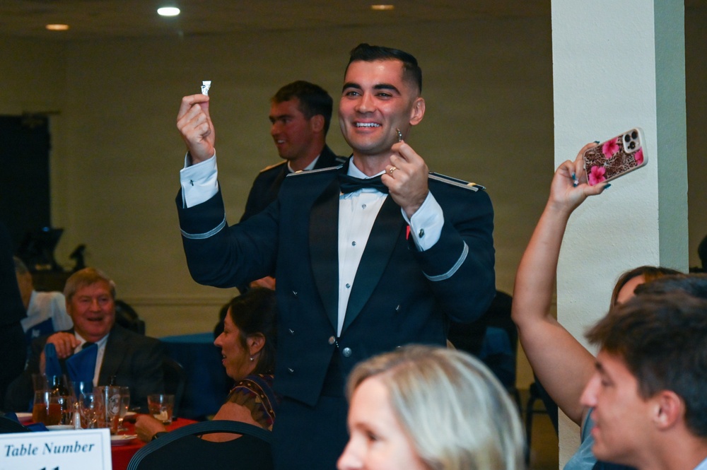 A formal military banquet or ball is taking place in a ballroom setting. A man in a dark military dress uniform with a bow tie is standing and smiling broadly, holding up what appears to be a small ticket or card in each hand, suggesting he may have won a raffle or prize. Guests seated at numbered tables are reacting, with one person raising a phone to take a photo.
