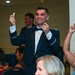 A formal military banquet or ball is taking place in a ballroom setting. A man in a dark military dress uniform with a bow tie is standing and smiling broadly, holding up what appears to be a small ticket or card in each hand, suggesting he may have won a raffle or prize. Guests seated at numbered tables are reacting, with one person raising a phone to take a photo.