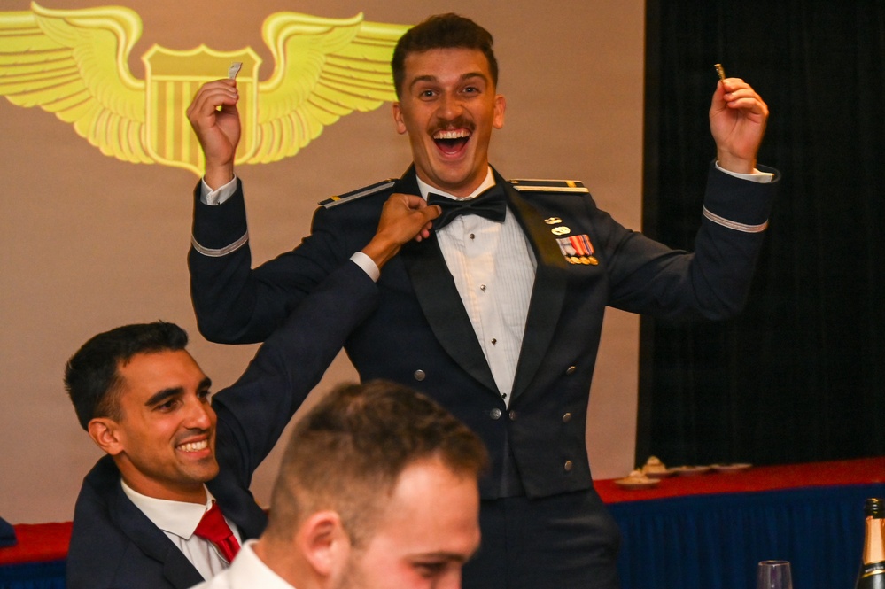 A US Air Force officer in dress mess uniform celebrates with arms raised and an exuberant expression during what appears to be a pilot wings ceremony or formal military dining event. A colleague behind him is pinning something onto his uniform, while other uniformed personnel are seated nearby. A projected aviator wings emblem is visible on the screen in the background.