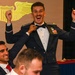 A US Air Force officer in dress mess uniform celebrates with arms raised and an exuberant expression during what appears to be a pilot wings ceremony or formal military dining event. A colleague behind him is pinning something onto his uniform, while other uniformed personnel are seated nearby. A projected aviator wings emblem is visible on the screen in the background.