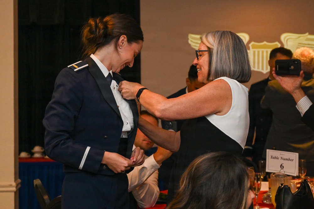 A formal military commissioning or promotion ceremony is taking place in a banquet hall setting. A woman in civilian attire is pinning insignia or a device onto the collar of a person wearing a dark blue military dress uniform. Both individuals are smiling, suggesting a celebratory and emotionally significant moment.